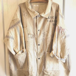 May include: Beige button-up shirt with vertical stripes and two front pockets. The shirt has rolled-up sleeves and text that reads "BLUE CANN DAY". The shirt is hanging on a wooden hanger.