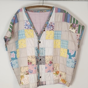 May include: A vintage patchwork vest with a button-down front. The vest features a variety of fabric squares in pastel colors, including floral and striped patterns. It hangs on a wooden hanger against a white wall.