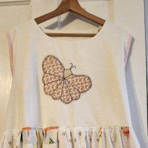 May include: White sleeveless dress with a butterfly applique. The butterfly is made of floral fabric and outlined in black. The dress has a patchwork skirt and pink trim along the neckline and sleeves. The dress is hanging on a wooden hanger.