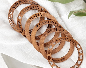 May include: Five wooden ring-shaped place cards with engraved names and dates. The light brown rings feature decorative cutouts of hearts and leaves. Names include Cameron, Maria, Michael, and Samantha.