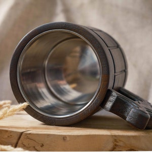 May include: A dark brown wooden beer stein with a stainless steel interior and a carved wooden handle. The stein is sitting on a wooden surface.