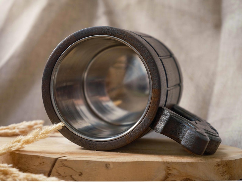 May include: A dark brown wooden beer stein with a stainless steel interior and a carved wooden handle. The stein is sitting on a wooden surface.