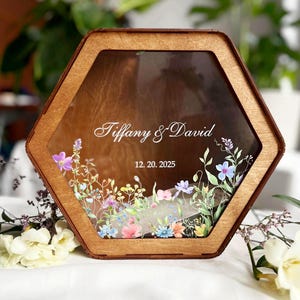 Könnte beinhalten: Eine sechseckige Holzbox mit einer transparenten Vorderseite, die unten mit einem bunten Blumenmuster bedruckt ist. Der Text "Tiffany & David" und das Datum "12. 20. 2025" sind in Weiß auf der transparenten Oberfläche der Box zu sehen.