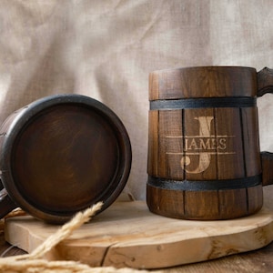 May include: Two wooden beer mugs with a dark brown finish. One mug has the name "JAMES" engraved on it. The mugs are sitting on a wooden surface.