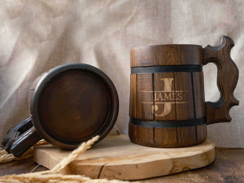 May include: Two wooden beer mugs with a dark brown finish. One mug has the name "JAMES" engraved on it. The mugs are sitting on a wooden surface.