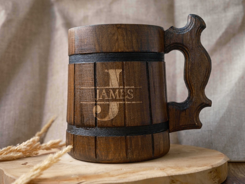 May include: A wooden beer stein with a dark brown finish. The stein has a handle and is engraved with the name "JAMES" and the letter "J".