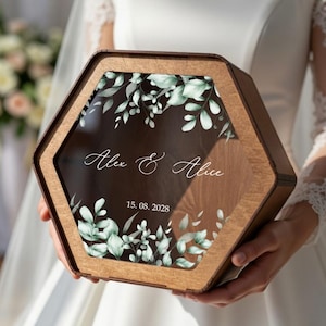 Può includere: Scatola esagonale in legno con piano in vetro trasparente. La scatola è incisa con i nomi "Alex & Alice" e la data "15. 08. 2028". Il vetro è decorato con elementi floreali verdi e bianchi.