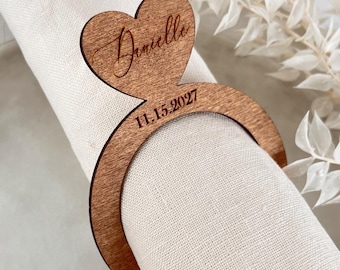 May include: A wooden napkin ring featuring a heart-shaped design with the name "Danielle" and the date "11.15.2027" engraved. The ring is placed on a rolled white napkin. The ring is made of wood.