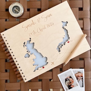 Może przedstawiać: Drewniany album podróżniczy z wyciętymi mapami Wielkiej Brytanii i Japonii, kompasem i dwoma zdjęciami. Album jest podpisany "Sarah & Sam 24th April 2026". Drewniany długopis spoczywa na stronie, z przerywaną linią i samolotem.