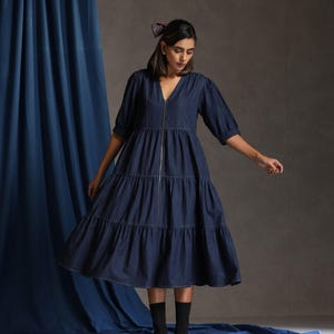May include: A dark blue denim dress with a V-neckline and a zip down the front. The dress has a tiered skirt and puffed sleeves. The model is wearing black ankle boots.