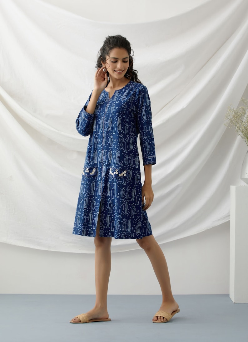 May include: A blue cotton dress with a white fish print and pearl embellishments on the pockets. The dress has a V-neck and three-quarter sleeves.