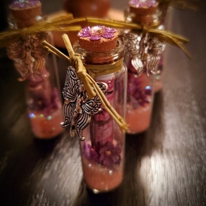 May include: Three small glass bottles with cork stoppers and a decorative butterfly charm. Each bottle is filled with pink glitter and purple flowers.