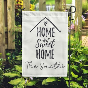 Peut inclure: Drapeau de jardin blanc avec du texte noir qui dit "Home Sweet Home The Smiths". Le drapeau est attaché à un poteau en métal noir et est affiché dans un jardin.