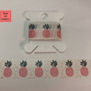 May include: A roll of white washi tape with a repeating pattern of pink pineapples and gold glitter. The tape is on a white spool.