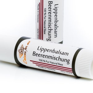 Könnte beinhalten: Zwei weiße Lippenbalsam-Tuben mit schwarzen und weinroten Akzenten. Die Tuben sind mit "Lippenbalsam Beerenmischung 100% Pur Natural Base" und "Made in Germany" beschriftet. Eine Tube liegt, die andere steht aufrecht.