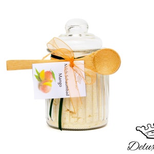 Könnte beinhalten: Ein klares Glas mit Deckel, gefüllt mit einem hellfarbigen Produkt. Ein Holzlöffel liegt auf dem Glas, gebunden mit einem orangefarbenen Band. Ein Etikett mit einer Mango-Illustration und dem Text "Milch-Schaumbad Mango" ist angebracht.