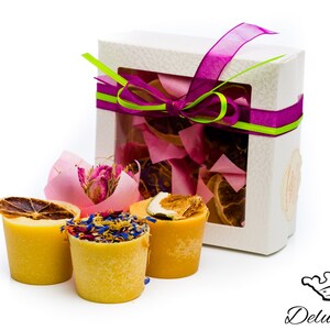 Può includere: Un set di quattro cere per profumare la casa di colore giallo in piccole tazze bianche, ciascuna con un diverso topping di fiori o frutta secca. Le cere sono confezionate in una scatola regalo bianca con un nastro viola e verde.