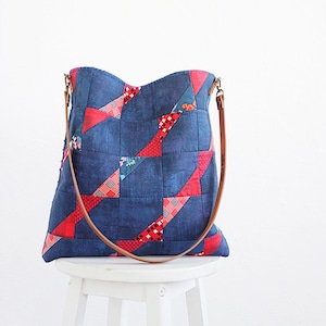 May include: A denim tote bag with a patchwork design featuring red and patterned fabric accents. The bag has brown leather straps and is displayed on a white stool. The text "ANNA STUDIO" is visible.