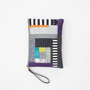 Könnte beinhalten: Eine Patchwork-Clutch mit einer Vielzahl von Stoffmustern und -farben, darunter Grau, Schwarz, Blau und Gelb. Die Clutch hat eine schwarze Handschlaufe und einen lila Rand. Der Text "ANNA STUDIO" befindet sich unten.