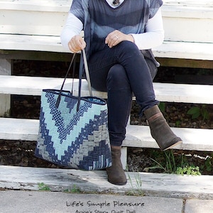 Może przedstawiać: Niebiesko-szara torba typu tote z geometrycznym wzorem, trzymana przez osobę. Torba ma ciemnoniebieskie skórzane wykończenia i uchwyty. Tekst "Life's Simple Pleasure" jest widoczny na dole obrazu.