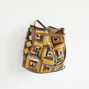 Puede incluir: Bolso tote patchwork hecho a mano con un patrón geométrico en tonos amarillos, marrones y negros. El bolso tiene asas de cuero marrón y una correa para el hombro. Un accesorio único y artesanal.