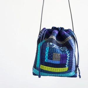 May include: A patchwork drawstring bag in shades of blue, purple, and teal. The bag features a variety of geometric patterns and textures, with a drawstring closure and dark blue straps. The bag is suspended against a white background.