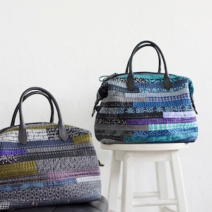 Puede incluir: Dos bolsos acolchados con asas de cuero negro. Los bolsos presentan un diseño de patchwork en tonos azules, morados, grises y verdes. Un bolso está sobre un taburete blanco. El texto "ANNA STUDIO" es visible.