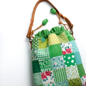 May include: A patchwork tote bag with a brown leather handle and green drawstring closure. The bag features a variety of green fabric squares with floral, polka dot, and plaid patterns. The bag has a white background.