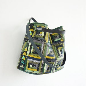 Pode incluir: Bolsa tote patchwork com um design geométrico em tons de verde, cinza e amarelo. A bolsa tem uma alça cinza escura e está pendurada em uma parede branca. O texto "ANNA STUDIO" é visível na parte inferior.