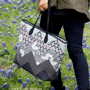 May include: A patterned tote bag with black handles is held by a person walking through a field of blue flowers. The bag features a geometric design in shades of gray, white, and teal. The text "Life's Simple Pleasure" is visible on the bag.
