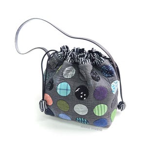 May include: A gray quilted drawstring pouch bag featuring various colorful fabric circles in shades of blue, green, orange, and purple. The bag has a black and white striped interior lining, a black strap, and the text ANNA STUDIO printed in white.