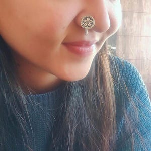 May include: A silver nose ring featuring a circular design with a floral pattern. The nose ring is worn on the nose. The jewelry is a small, decorative accessory.