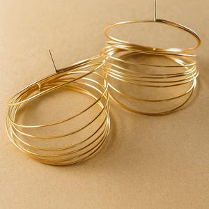 May include: A pair of gold-toned earrings constructed from multiple thin, circular wire loops stacked together. Each earring features a central vertical post attached to the top of the layered wire structure, creating a multi-dimensional hoop design.