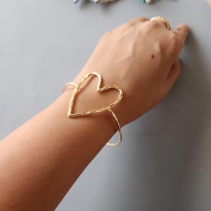 Può includere: Un bracciale a cuore aperto color oro. Il bracciale presenta una texture martellata e un design semplice ed elegante. La forma a cuore è il punto focale, con una fascia delicata che avvolge il polso. Gioielli.