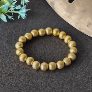 Op de afbeelding: Een cirkelvormige armband bestaande uit ronde, getextureerde goudkleurige kralen. De kralen hebben een matte, korrelige oppervlakteafwerking en zijn aan elkaar geregen in een doorlopende lus, wat zorgt voor een uniforme uitstraling van het sieraad.