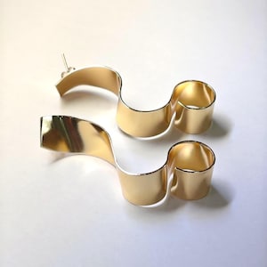 May include: A pair of gold-toned earrings featuring a smooth, continuous ribbon-like design that curves into an S-shape. Each earring has a polished, reflective surface and includes a thin metal post backing for attachment to the earlobe.