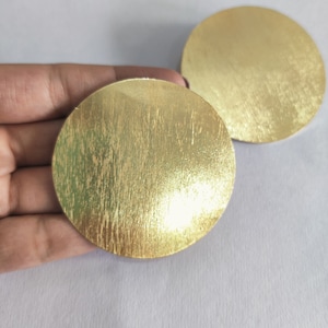 Puede incluir: Dos discos grandes y redondos de metal dorado con una superficie texturizada. Los discos se muestran sobre un fondo blanco. Los discos son probablemente componentes para joyería u otros proyectos de artesanía.