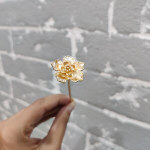 May include: A gold flower hair pin with a delicate design. The pin is held in a hand against a white brick background.