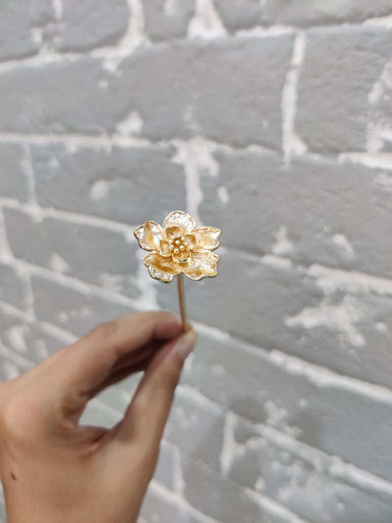 May include: A gold flower hair pin with a delicate design. The pin is held in a hand against a white brick background.