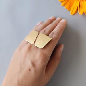 May include: A gold-coloured ring featuring two large, geometric shapes. The ring is worn on a hand, set against a grey background with a bright orange flower in the corner.