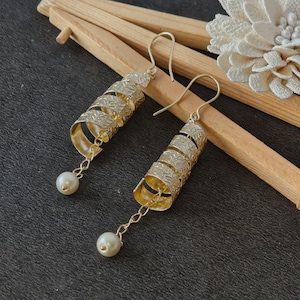 May include: A pair of gold-colored spiral earrings with a textured design. Each earring has a small, dangling pearl at the bottom. The earrings are displayed on a dark surface with wooden accents and a white floral embellishment.