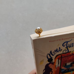 Op de afbeelding: Een goudkleurige metalen bladwijzer met een parel aan de bovenkant is in een boek gestoken. De bladwijzer heeft een bloemenontwerp. De kaft van het boek is zichtbaar, met de titel "Mrs. Funny" en de naam van de auteur.