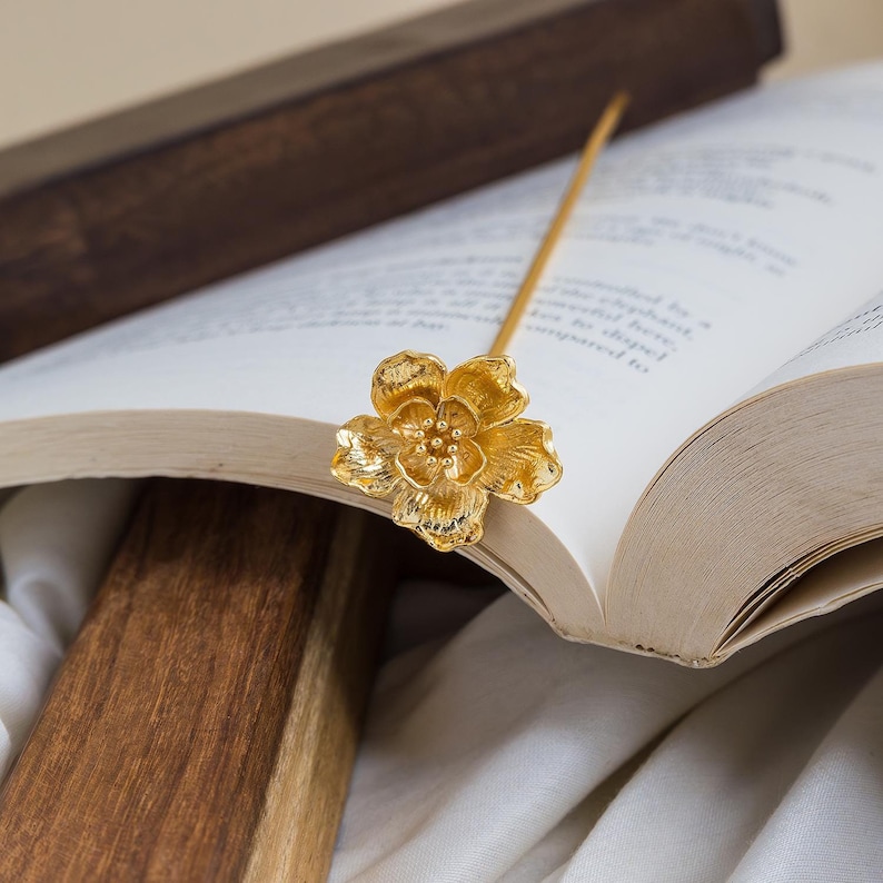 A detailed gold flower bookmark with a long, thin stem. The bookmark is placed in the pages of a closed book.