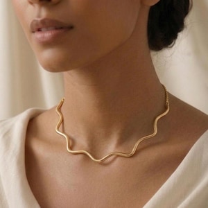 Puede incluir: Un collar de tono dorado que presenta un diseño de barra de metal lisa y continua con forma ondulada o serpentina que descansa sobre el cuello. La pieza está conectada a una cadena delicada en la parte posterior, con un acabado metálico.