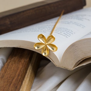Könnte beinhalten: Ein goldfarbenes Blumen-Lesezeichen mit einem langen, dünnen Stiel, das in die Seiten eines geöffneten Buches gelegt wird.