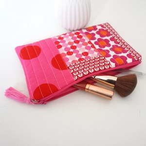 Könnte beinhalten: Eine gesteppte Stofftasche mit einem Patchwork-Design aus rosa und roten Punkten, Blumenmustern und geometrischen Formen. Die Reißverschlusstasche hat einen rosa Quastenanhänger und enthält einen Make-up-Pinsel sowie einen Lippenstift.