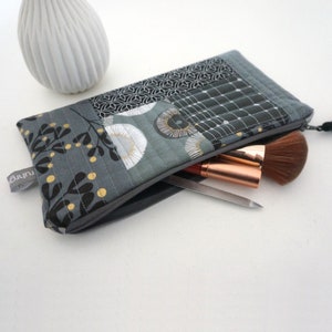 Könnte beinhalten: Eine gesteppte Kosmetiktasche mit grauem und schwarzem Blumenmuster, teilweise geöffnet, um Make-up-Pinsel und eine Pinzette zu zeigen. Die Tasche hat einen Reißverschluss und ein kleines Etikett mit dem Text "ruhrg".