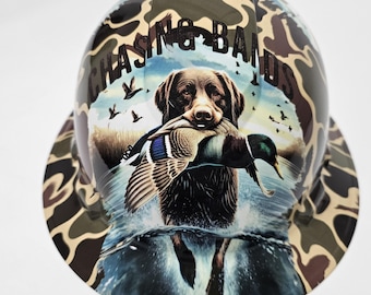 Puede incluir: Un casco de seguridad de camuflaje con un perro marrón que lleva un pato en la boca. El casco tiene las palabras "Chasing Bands" impresas.