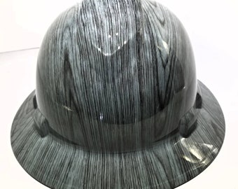 Puede incluir: Un casco gris con un patrón de veta de madera. El casco tiene una visera y un sistema de suspensión.