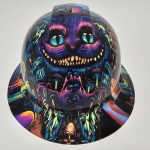 May include: A hard hat with a vibrant, psychedelic design featuring the Cheshire Cat from Alice in Wonderland. The hat is predominantly black with purple, blue, and green accents. Text on the hat reads "we're all mad here."